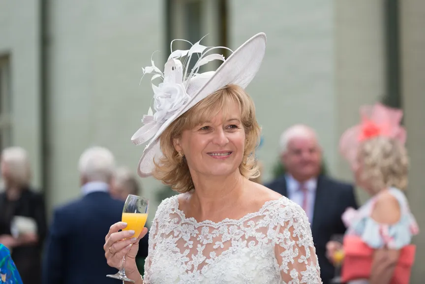 Smiling woman in a white lace dress and large white fascinator hat holding a glass of orange juice at a social event.