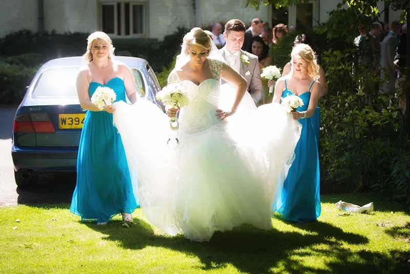 Bride in a white wedding dress walking outdoors with two bridesmaids in blue dresses holding her dress train, and a groom behind her.