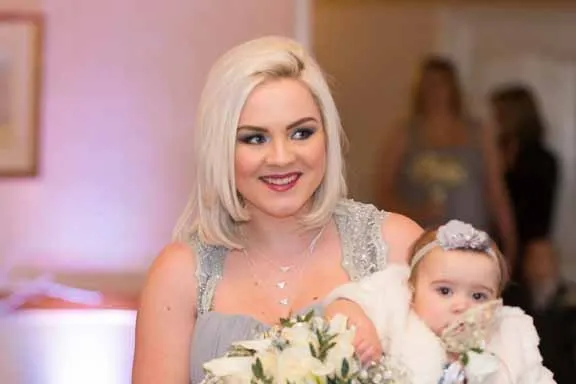 Blonde woman in a silver dress holding a baby girl dressed in white with a headband and flower accessory.