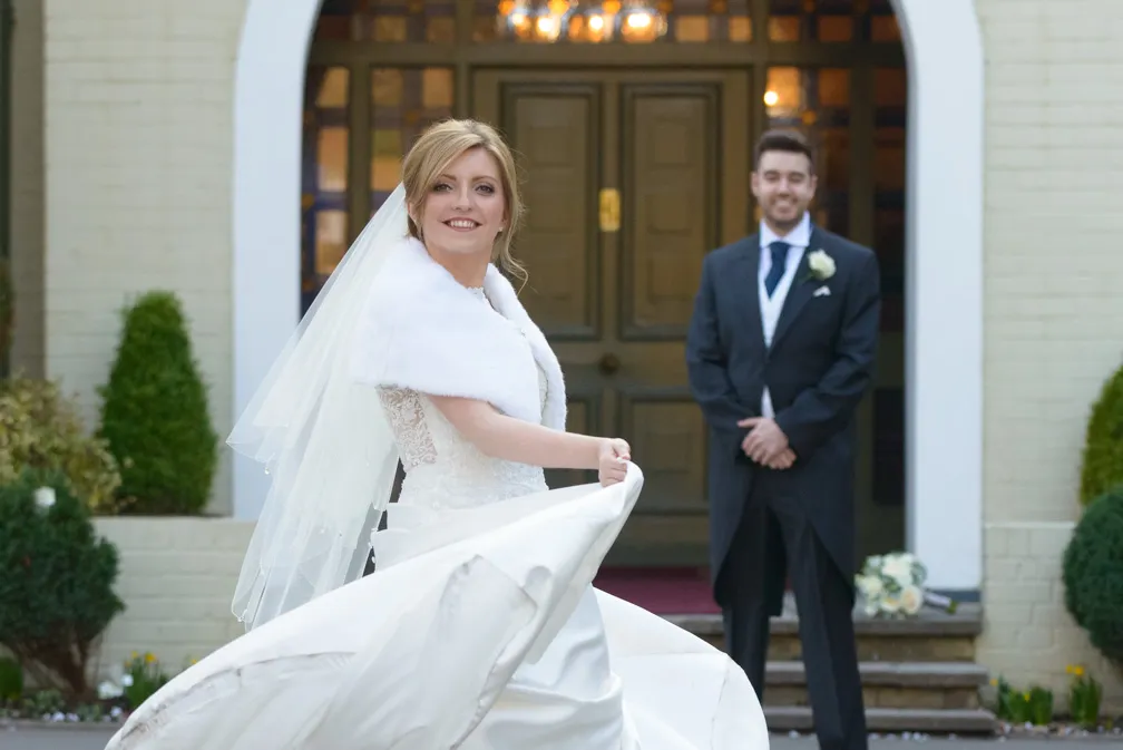 Bride twirling in a white wedding dress and veil outdoors, with a groom in a dark suit smiling in the background.