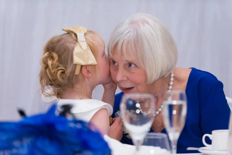 Young girl whispering a secret to an elderly woman wearing a blue dress and pearl necklace at a table.