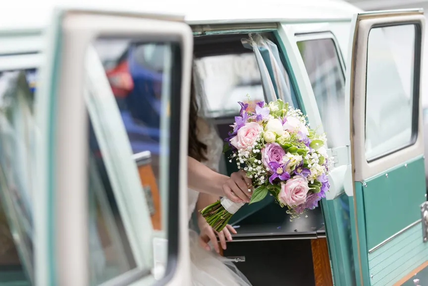 Bride holding a bouquet of pink, purple, and white flowers while seated inside a green vintage vehicle with the door open.