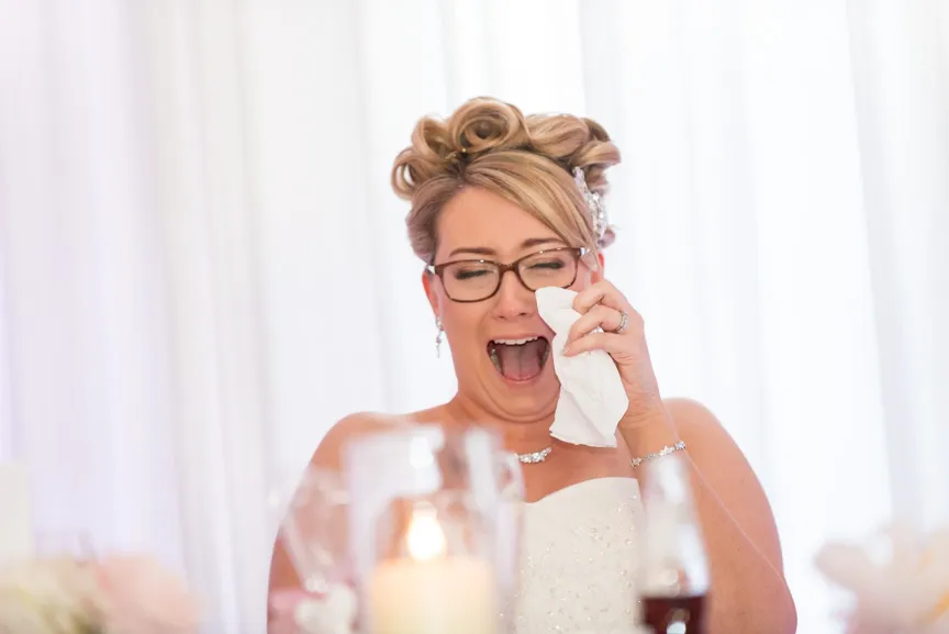 Bride in wedding dress and glasses wiping tears with tissue during emotional moment.