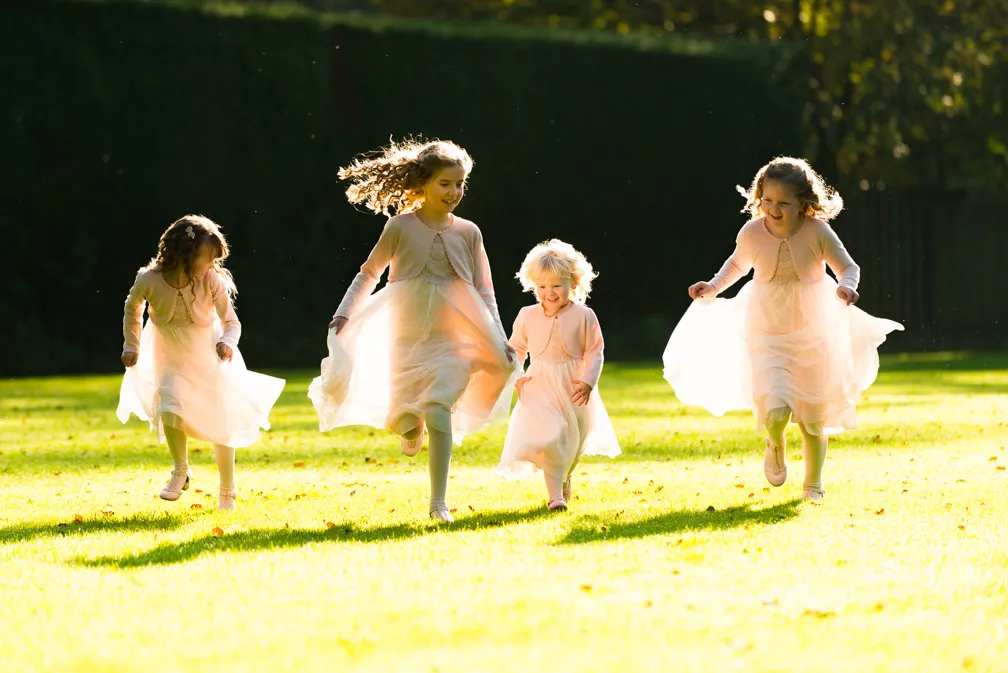 Four young girls in light pink dresses running and playing on a sunlit grassy lawn.