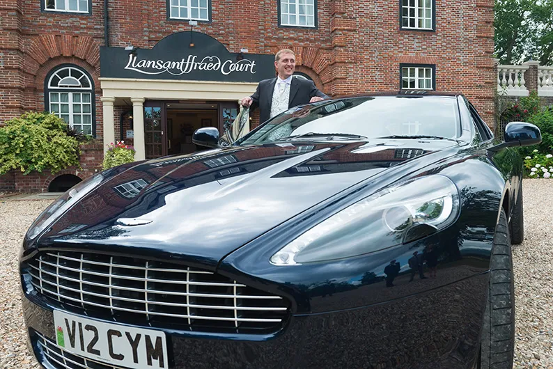 Man in a suit standing next to a dark blue luxury sports car with license plate V12 CYM in front of a brick building with a sign reading Llansantffraed Court.