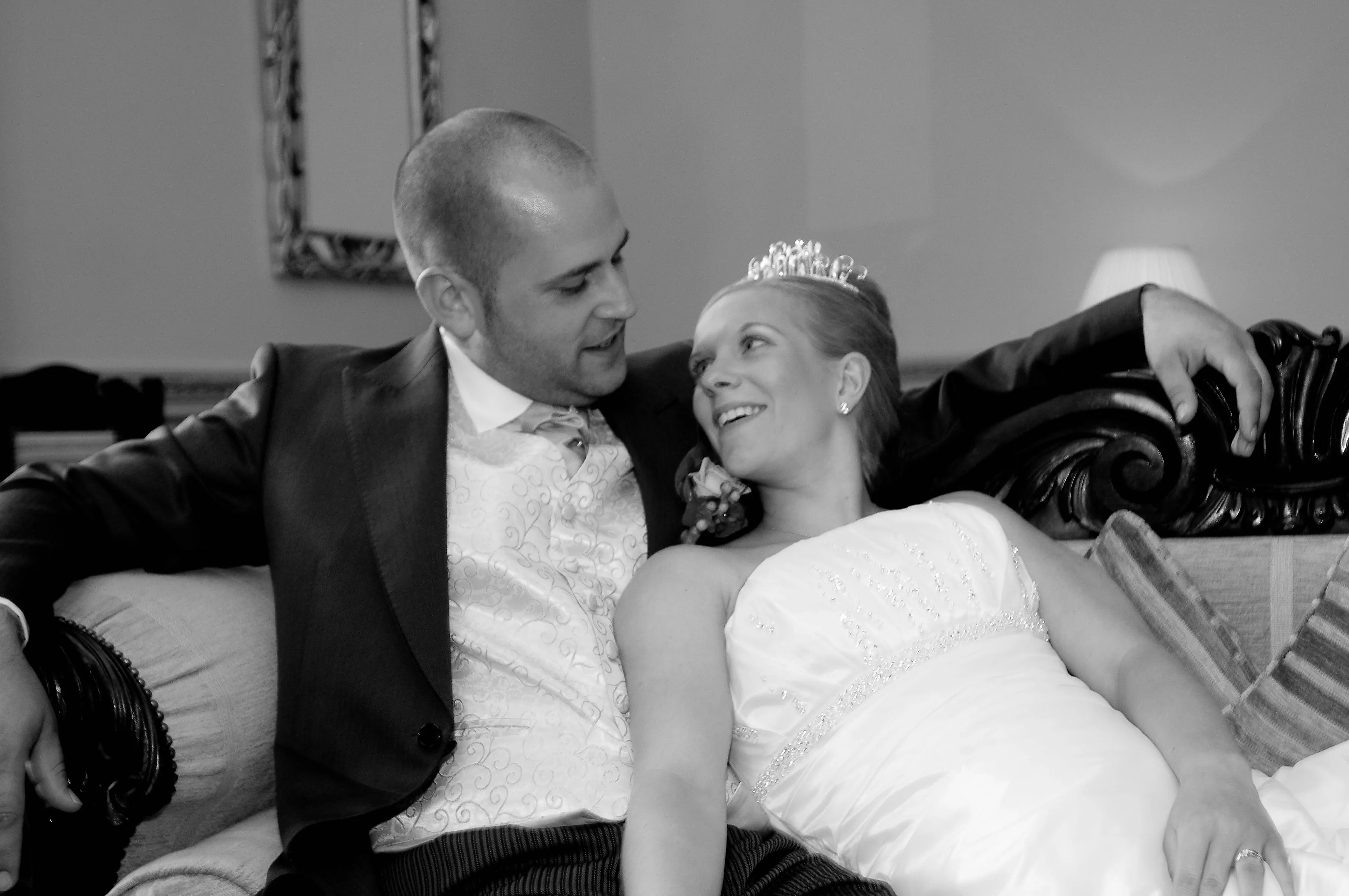 Bride in wedding dress and tiara leaning on groom in suit and vest, sitting closely on a couch looking at each other and smiling.