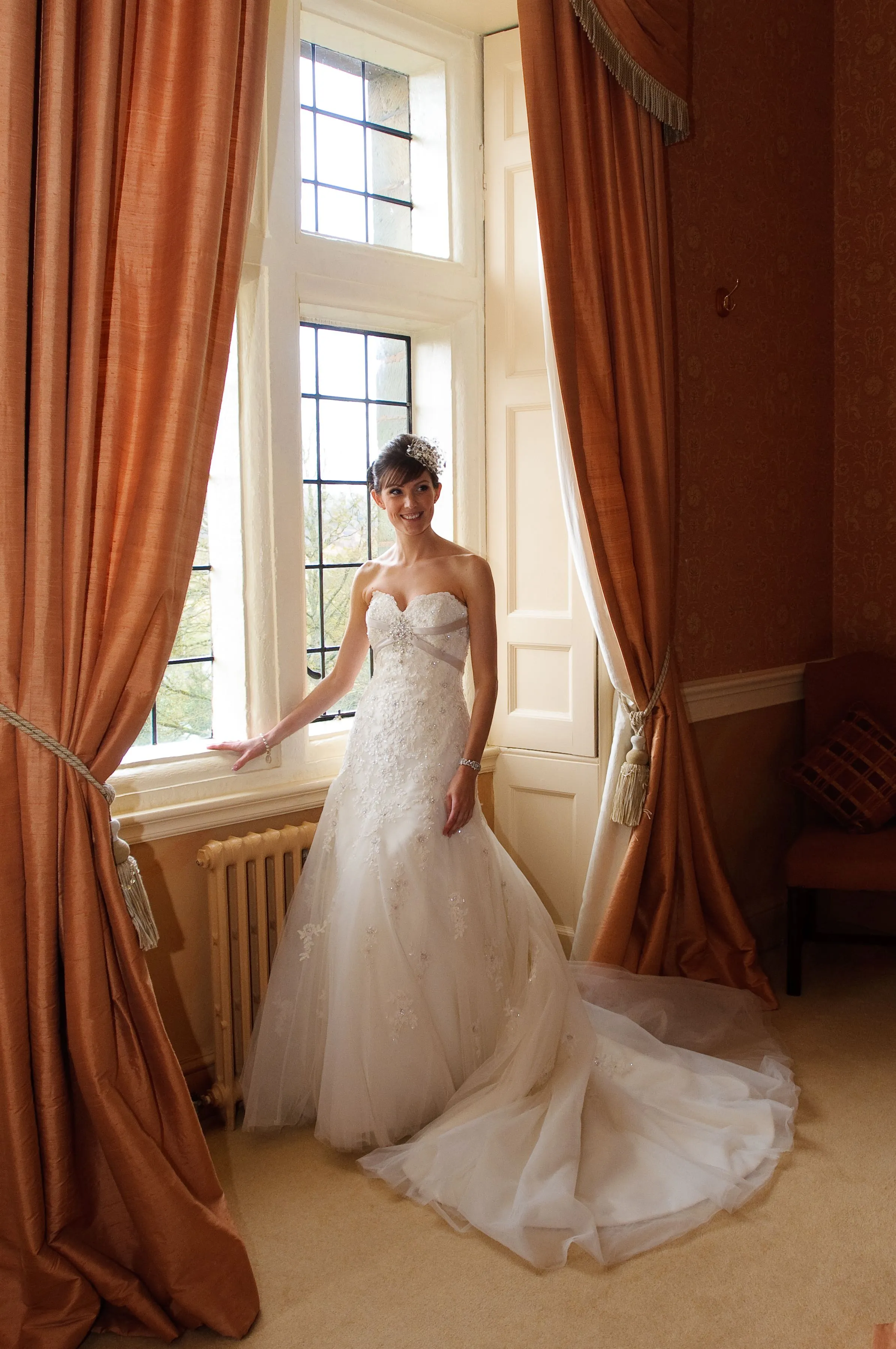 Bride in a strapless white wedding gown with floral details standing by a window with large orange curtains.