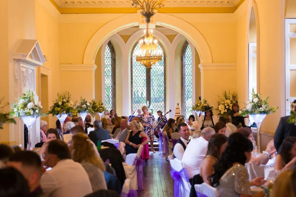 Guests seated at decorated tables with floral centerpieces and purple ribbons in a bright room with chandeliers and large leaded glass windows during a formal event.
