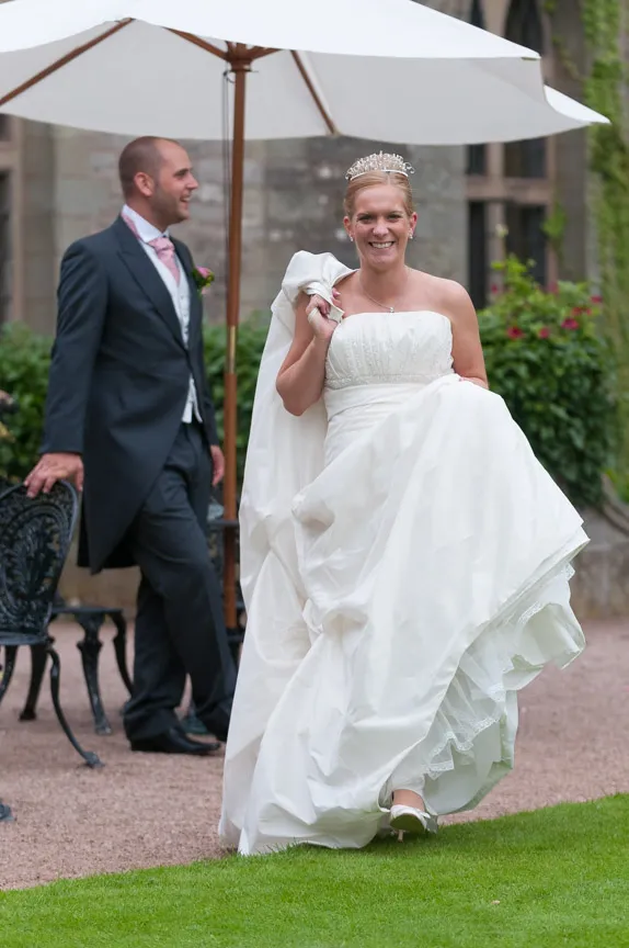 Bride in white dress and tiara smiling while lifting her gown, groom in suit standing behind her near an umbrella and garden chairs.