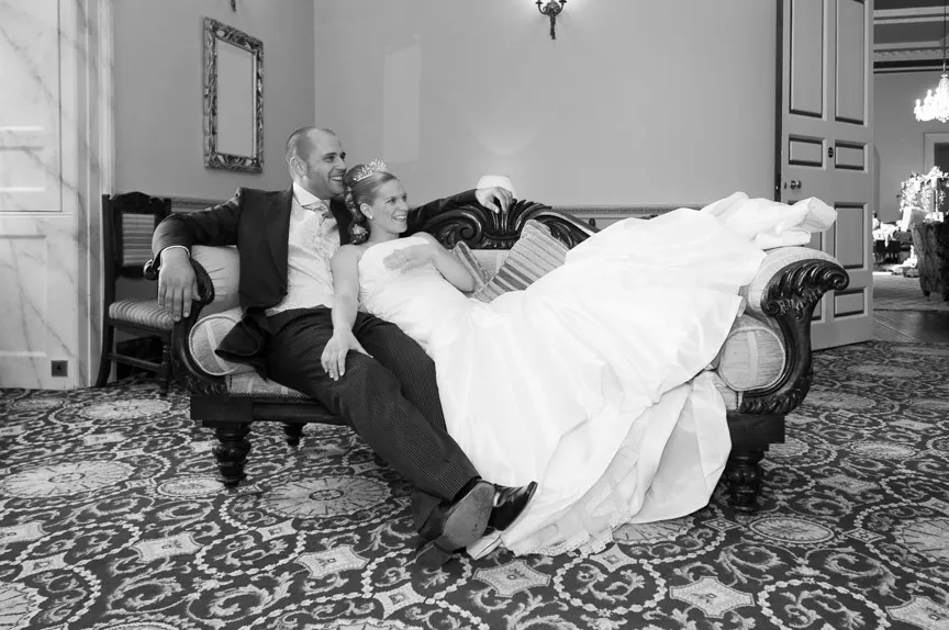 Bride and groom relaxing on an ornate sofa in a decorated room, bride wearing a wedding dress and groom in a suit.