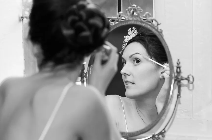 Woman applying makeup on her eye while looking into an ornate oval mirror.