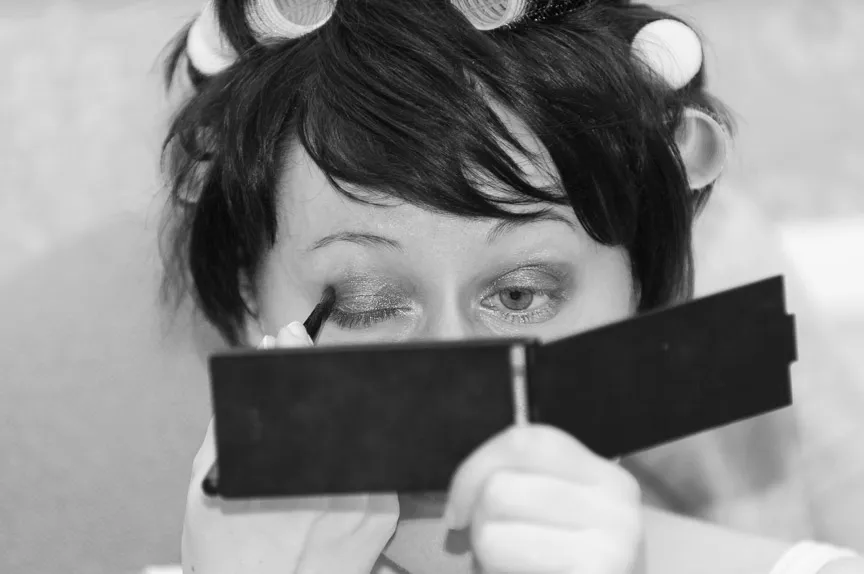 Woman with hair rollers applying eyeshadow while looking into a small mirror.
