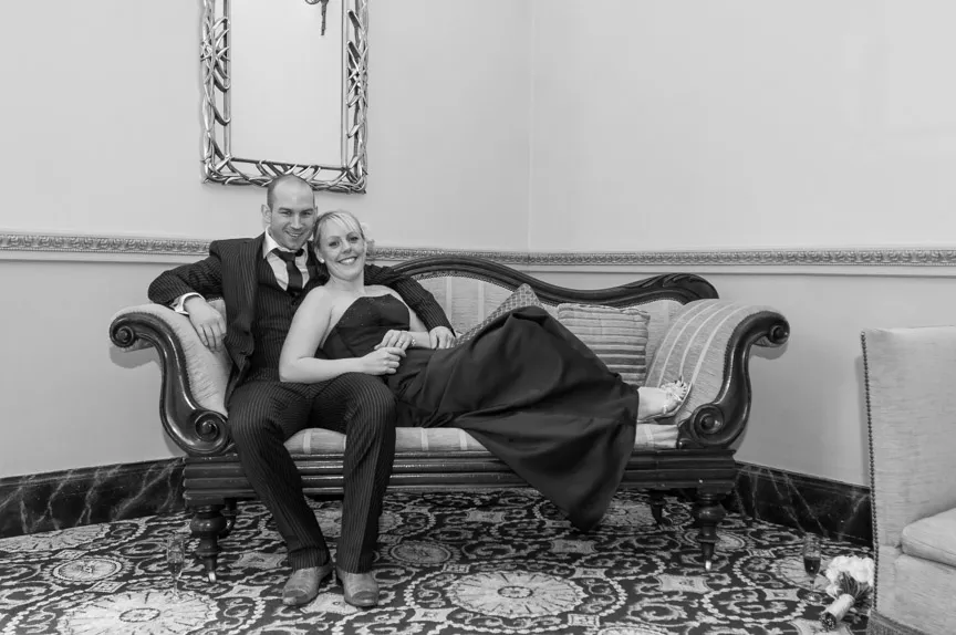 Couple dressed formally sitting on a vintage sofa with the woman reclining across the man's lap in a room with patterned carpet and ornate mirror.