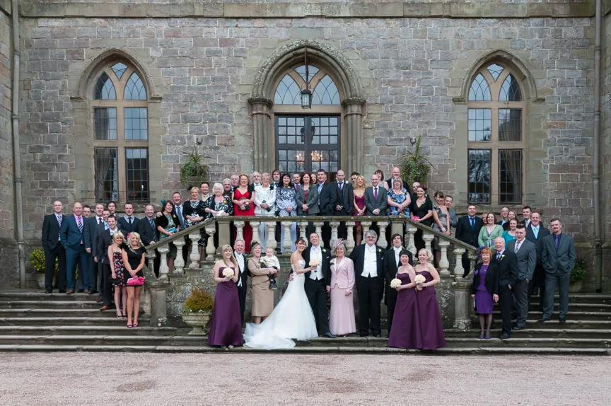 Large wedding group posed on stone steps in front of a historic building with arched windows, featuring bride in white gown and groom in tuxedo at the center.