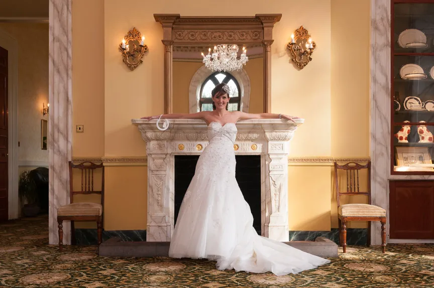 Bride in a strapless white wedding gown standing with arms outstretched on a marble fireplace mantel in an elegant room with ornate lighting and a chandelier.