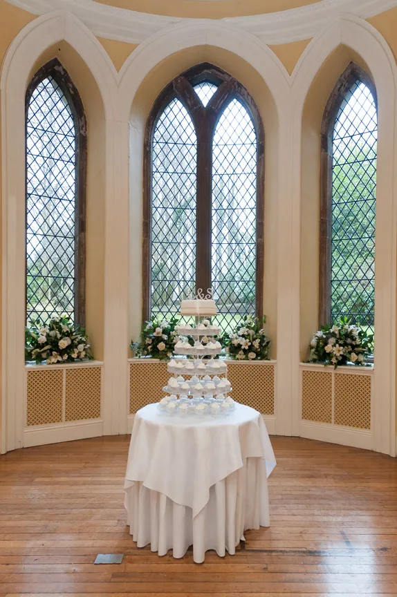 Tiered wedding cake with cupcakes on a round table with white tablecloth in front of large arched windows with floral arrangements on ledges.