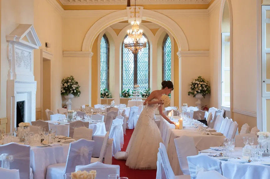 Bride in a white wedding dress lighting candles on a decorated table in an elegant reception room with white chair covers and floral arrangements.