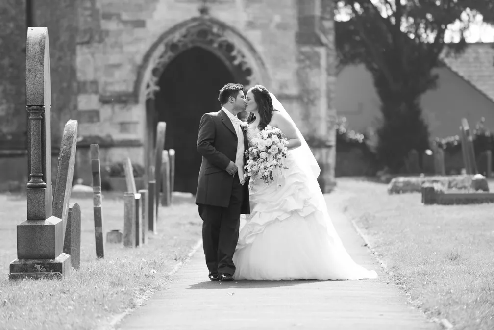 Bride and groom kissing on a path in a cemetery with an arched church entrance in the background, bride holding a bouquet.