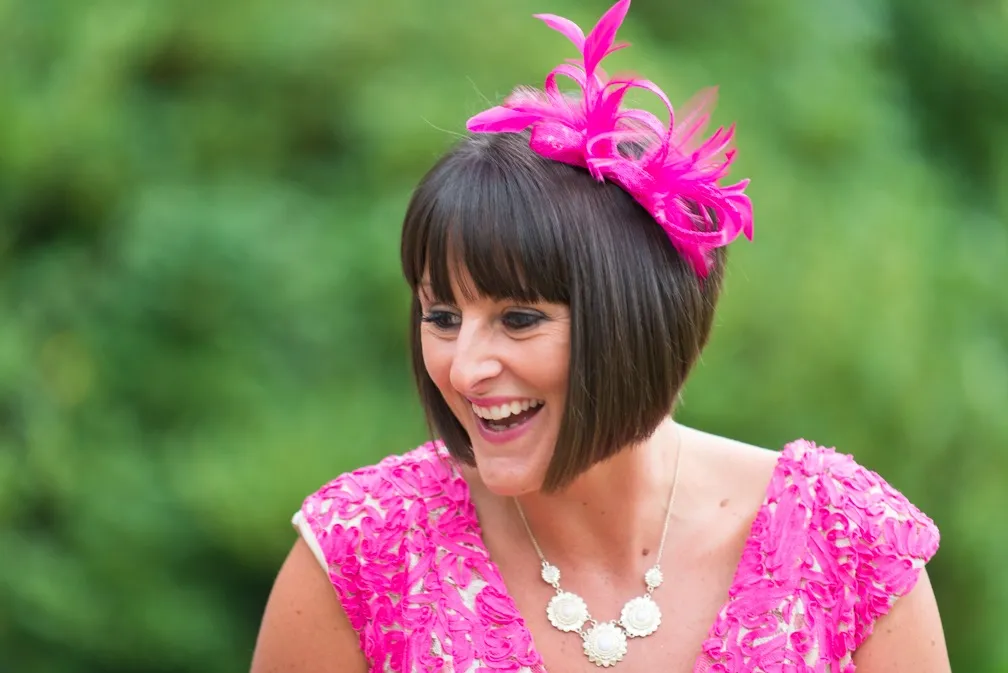 Smiling woman with short brown hair wearing a bright pink lace dress and a matching pink feathered fascinator headpiece.