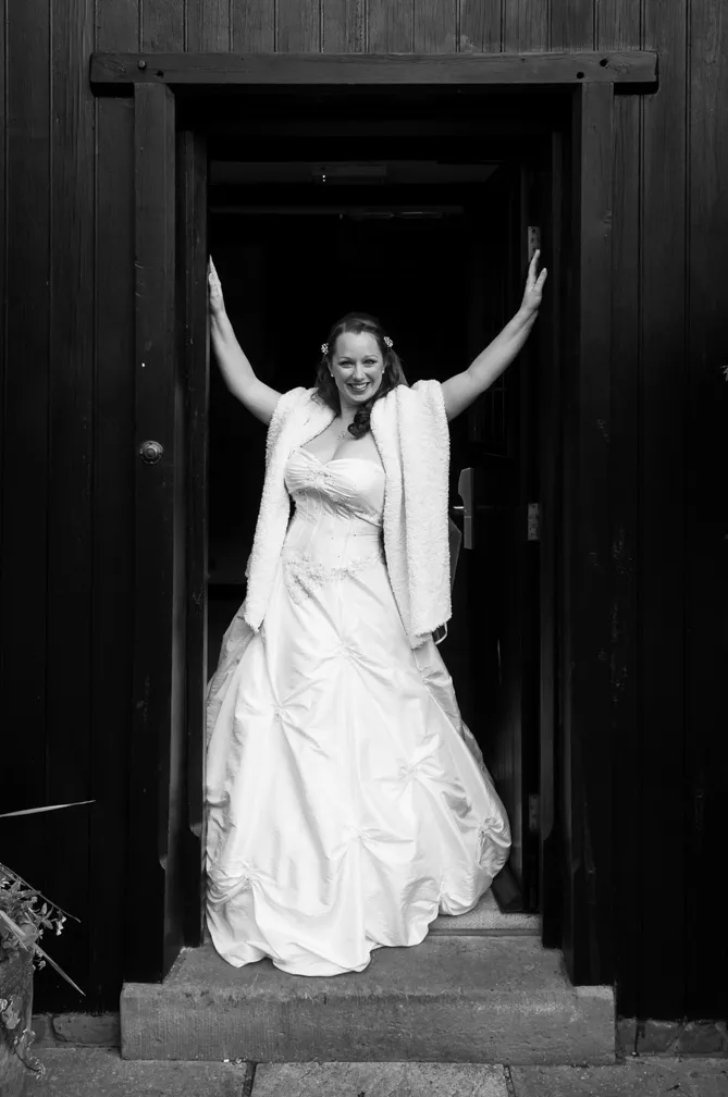 Bride in a white wedding gown and shawl standing in a dark doorway with arms raised and smiling.