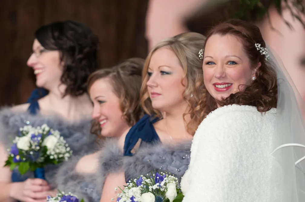 Bride in white shawl smiling at the camera with three bridesmaids in blue dresses holding bouquets.