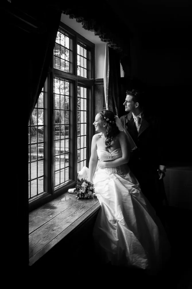 Bride in wedding dress holding bouquet and groom in suit standing by a large window looking outside, black and white photo.