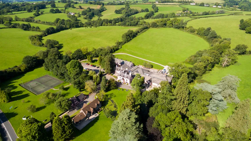 Aerial view of a countryside estate with a large house surrounded by trees, tennis courts, and green fields.