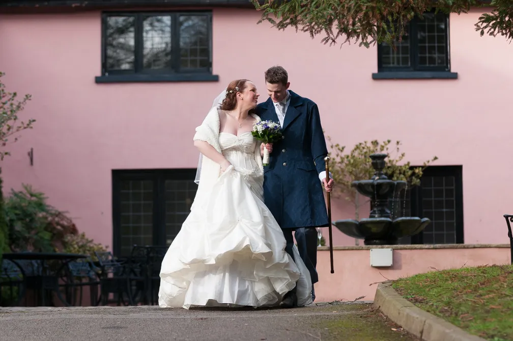 Bride in white gown holding a bouquet and groom in navy coat walking together outdoors near a pink building with black windows and a water fountain.