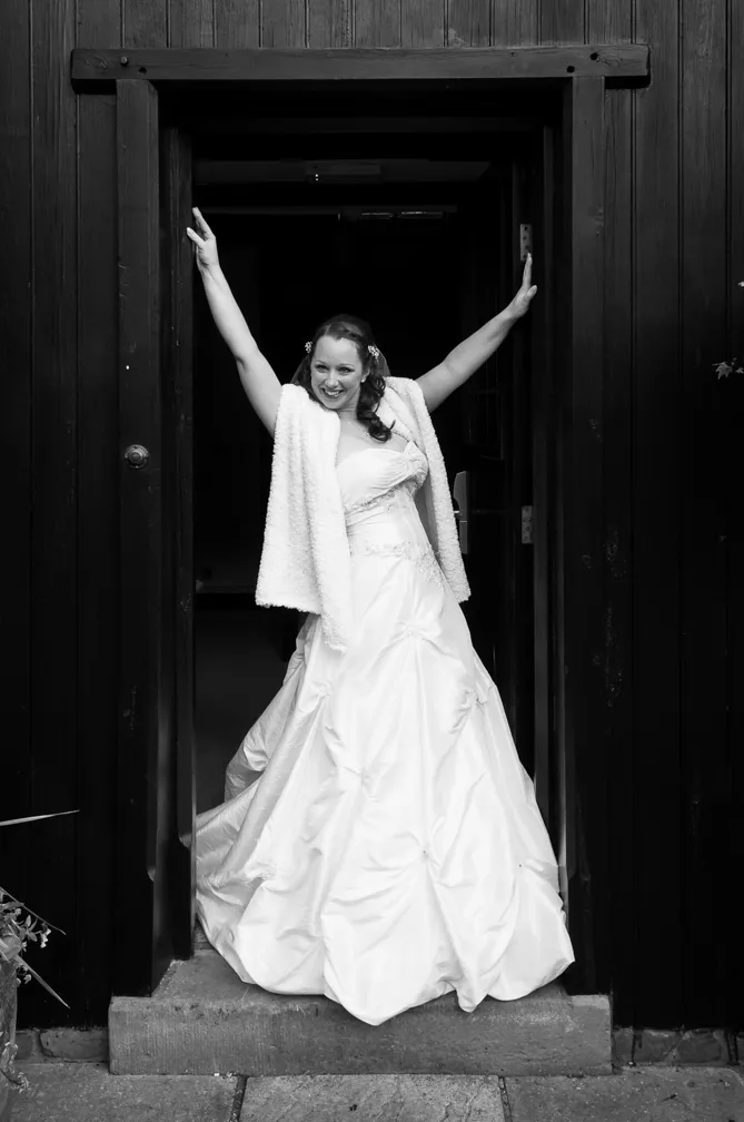 Smiling bride in a strapless wedding gown and shawl standing in a dark wooden doorway with arms raised.
