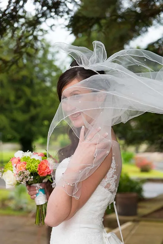 Smiling bride in a lace wedding dress holding a bouquet with a sheer veil blowing across her face outdoors.