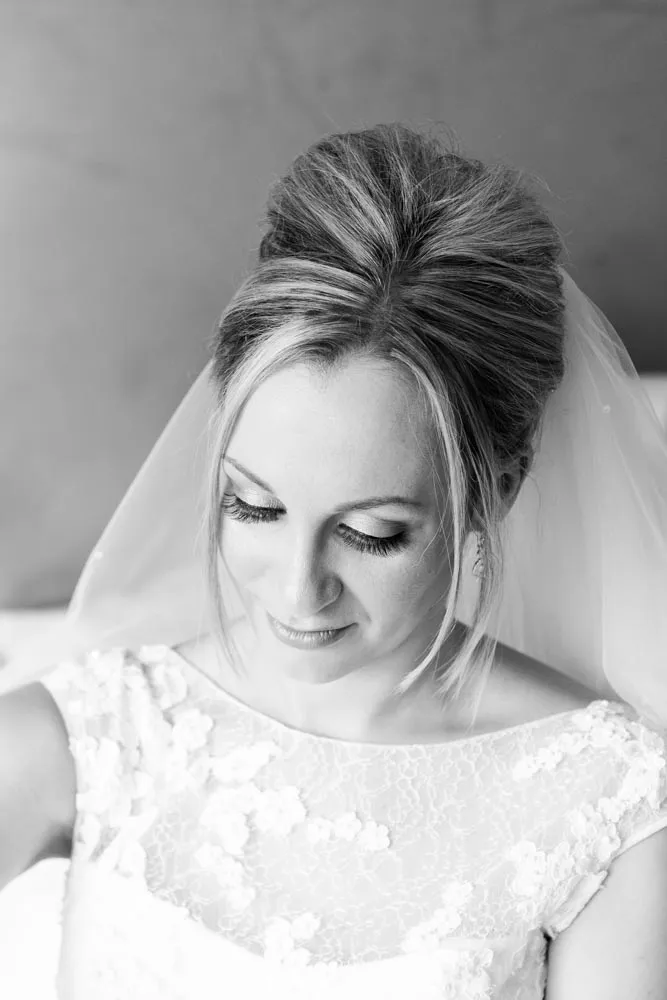 Black and white portrait of a bride with an elegant updo hairstyle, veil, and lace wedding dress looking down and smiling softly.