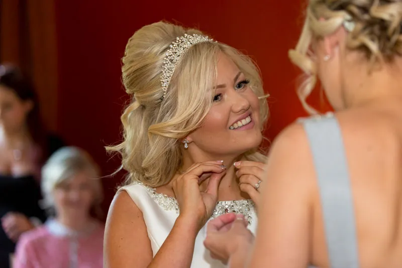 Bride with blonde hair wearing a jeweled headband and white dress, smiling while putting on earrings.