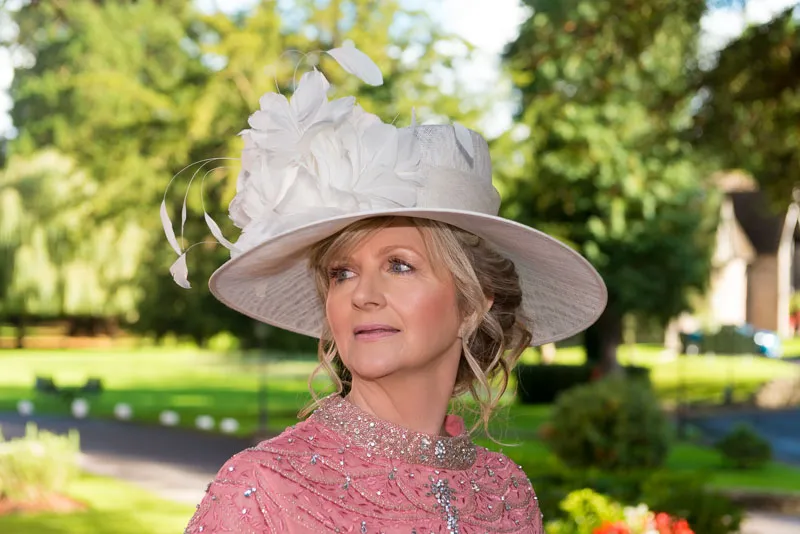Middle-aged woman wearing an elegant white hat adorned with large flowers and a pink embellished shawl, outdoors in a garden setting.