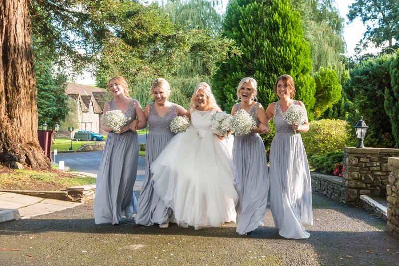 Bride in white wedding dress laughing and walking outdoors arm-in-arm with four bridesmaids in matching light gray dresses holding bouquets of white flowers.
