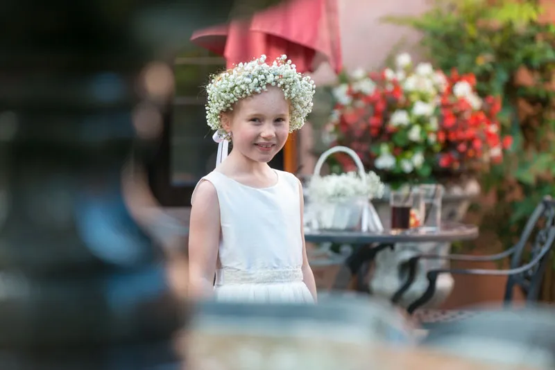 Smiling young girl wearing a white dress and a garland of white flowers on her head, standing outdoors near a table with drinks and potted flowers.