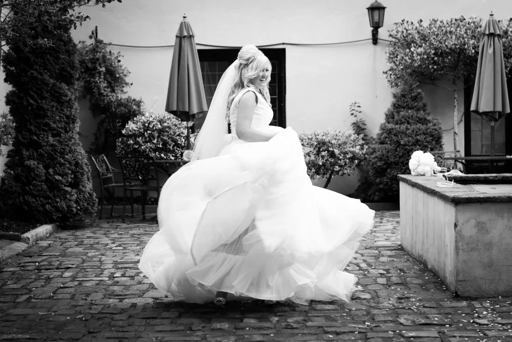 Bride spinning in a wedding dress with veil on a cobblestone courtyard surrounded by trees and umbrellas.