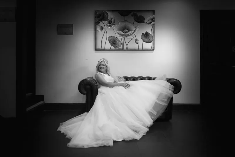 Bride in a flowing wedding gown reclining on a dark leather sofa under a floral painting.