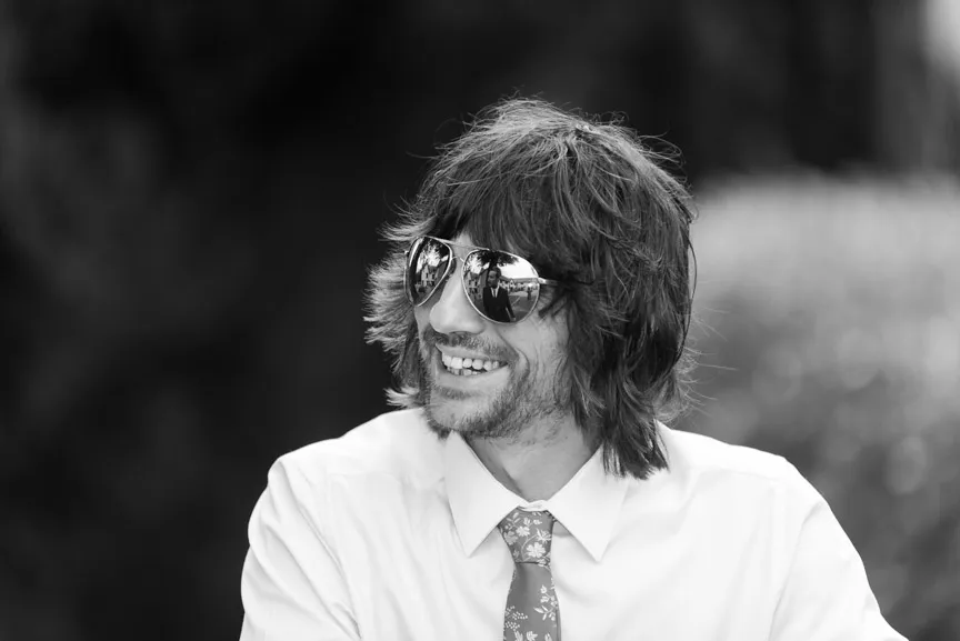 Smiling man with shaggy hair wearing reflective sunglasses, a white shirt, and a floral-patterned tie.
