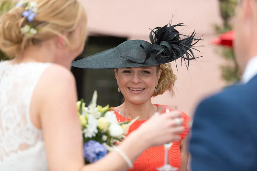 Woman in a bright orange dress and large black feathered hat smiling and holding a glass during an outdoor event.
