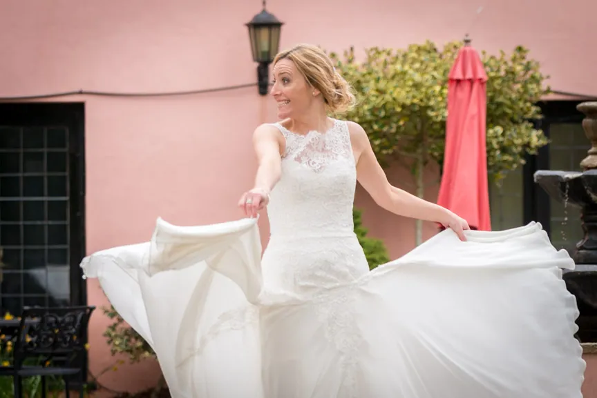 Smiling bride in a white lace wedding dress twirling outdoors in front of a peach-colored wall.