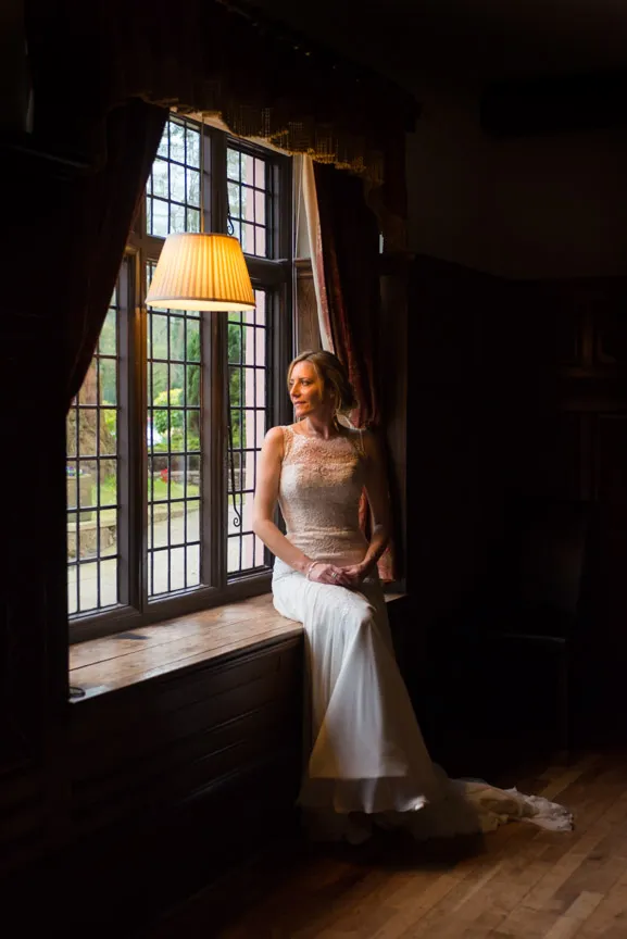 Bride in a lace wedding dress sitting on a wooden window bench with light from a window lamp illuminating her.