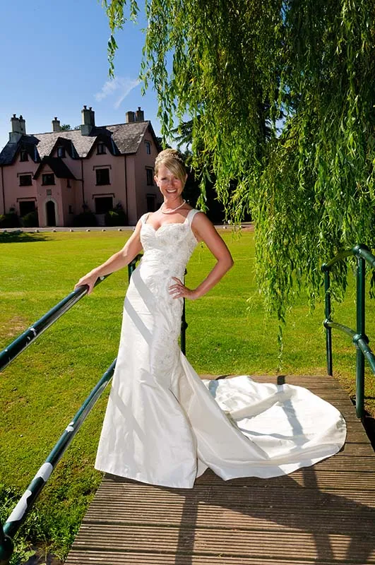 Smiling bride in a white wedding dress standing on a small wooden bridge under hanging tree branches with a house in the background.
