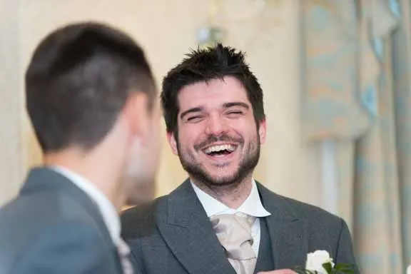Two men in suits smiling and talking, one facing the camera and laughing.
