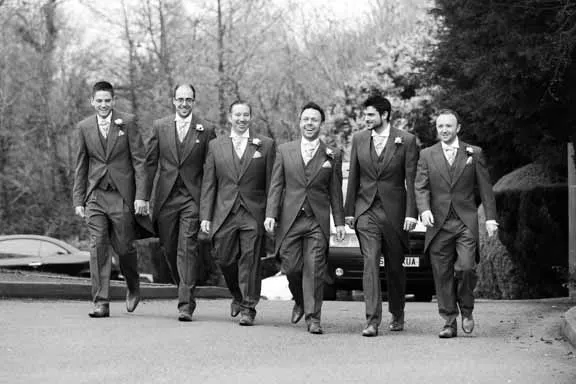 Six men wearing matching suits with boutonnieres walking together outdoors on a paved path.