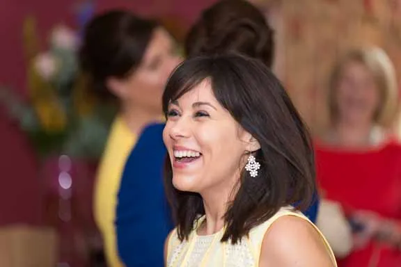 Smiling woman with dark hair and earrings in a social setting with blurred people in the background.