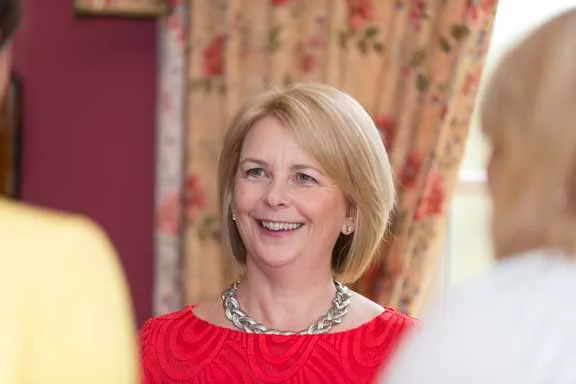 Smiling middle-aged woman with blonde hair wearing a red top and silver chain necklace, speaking with two blurred individuals.