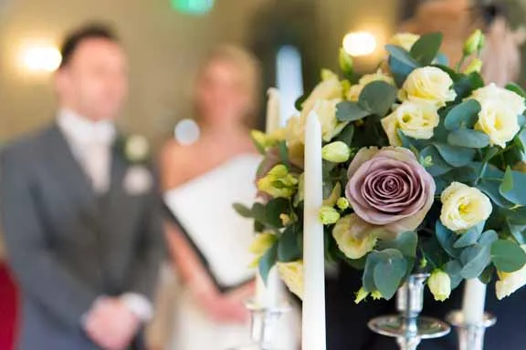Wedding ceremony scene with a floral arrangement of white and purple roses and green leaves in the foreground and a bride and groom blurred in the background.