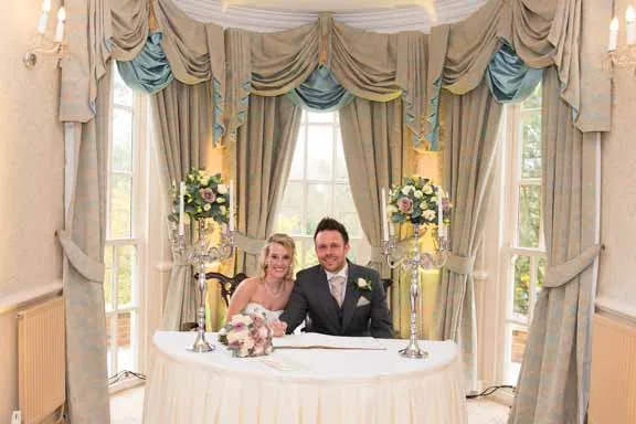Bride and groom seated at a table, smiling during a wedding ceremony in an elegant room with large windows and draped curtains.