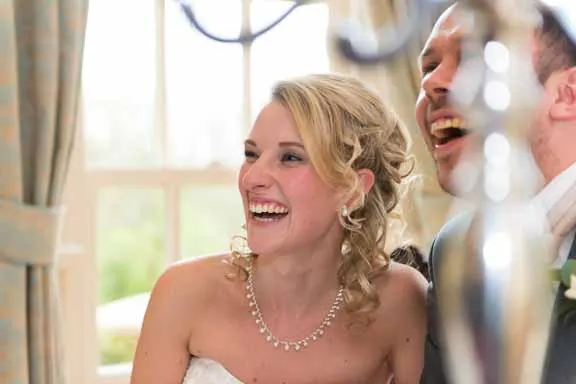 Bride with curly hair and necklace laughing beside groom during a wedding celebration.