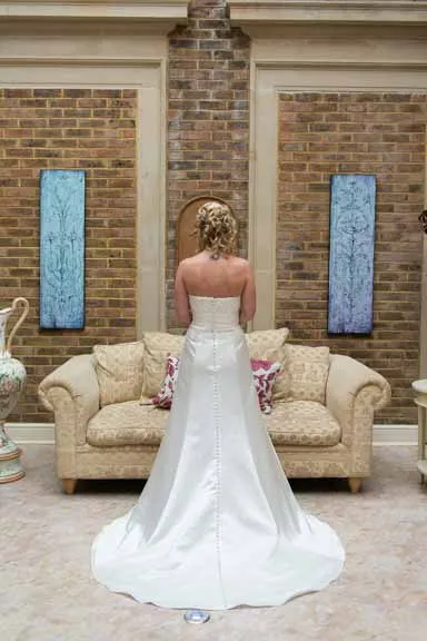 Bride in a strapless white wedding gown standing with her back to the camera in front of a beige sofa against a brick wall with two vertical blue decorative panels.